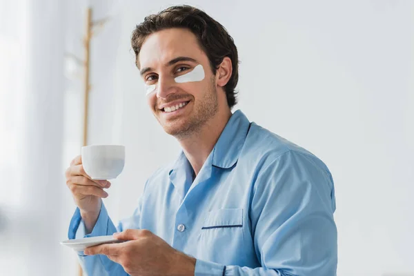 Giovane uomo in pigiama e patch per gli occhi che tiene il caffè e sorride alla fotocamera a casa — Foto stock