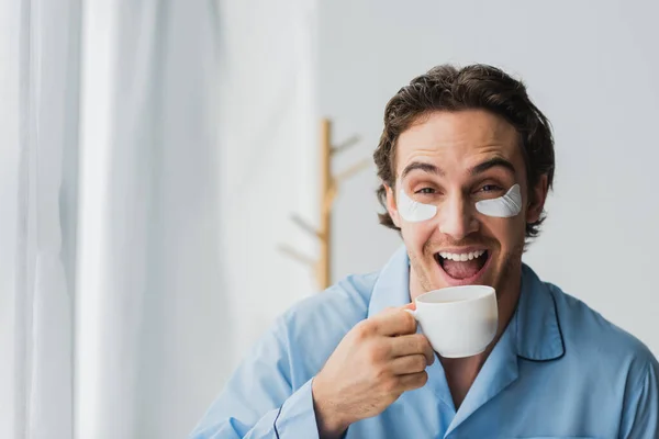 Homem alegre em manchas nos olhos e pijama segurando xícara de café em casa — Fotografia de Stock