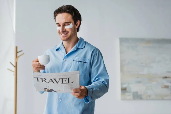 Uomo sorridente con toppe agli occhi e pigiama che tiene il caffè e legge il giornale di viaggio a casa — Foto stock