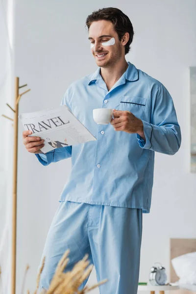 Joven con parches en los ojos y pijama sonriendo mientras lee noticias de viajes y sostiene café en el dormitorio - foto de stock