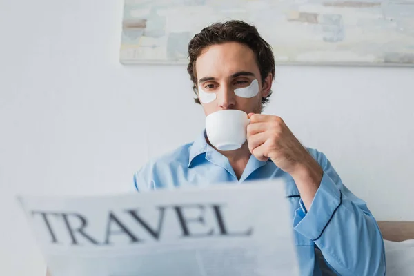 Giovane uomo con toppe agli occhi e pigiama che beve caffè e legge giornali sfocati in camera da letto — Foto stock