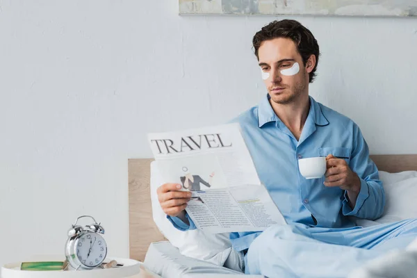 Hombre con parches en los ojos sosteniendo café y leyendo el periódico en la cama por la mañana - foto de stock