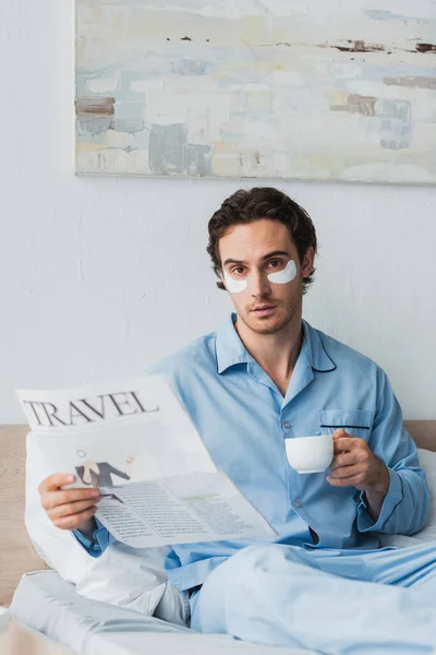 Young man with eye patches holding cup of coffee and travel news on bed at home — Stock Photo