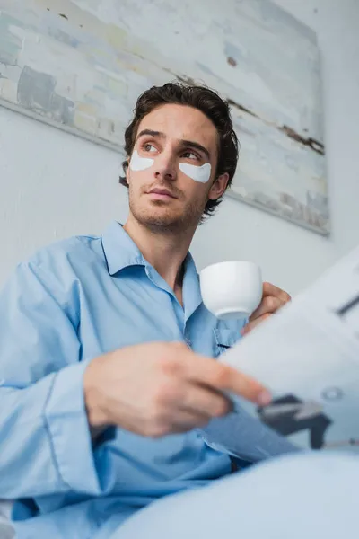 Vista de bajo ángulo del hombre en parches para los ojos y pijama con taza de café y periódico borroso en el dormitorio - foto de stock