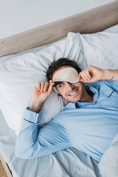 Высокий угол обзора улыбающегося человека в маске для сна и пижаме, лежащих на кровати утром — стоковое фото