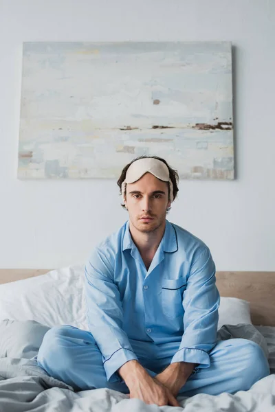 Joven con máscara de dormir y pijama sentado en la cama y mirando a la cámara - foto de stock