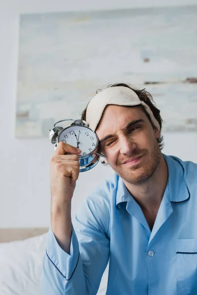 Irritated man in pajama holding alarm clock on bed at home — Stock Photo