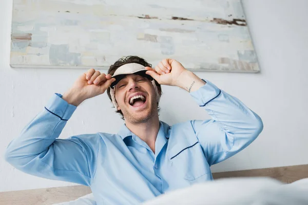 Vista a basso angolo di uomo positivo in maschera di sonno e pigiama guardando la fotocamera sul letto sfocato a casa — Foto stock