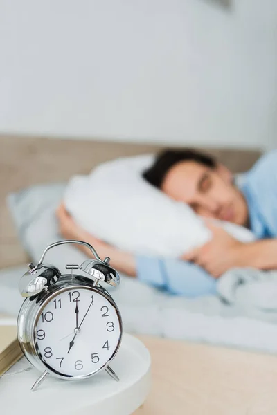 Reloj despertador y libro en la mesita de noche cerca del hombre borroso durmiendo en la cama - foto de stock