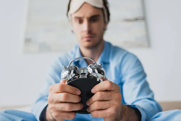 Alarm clock in hands of blurred man in sleep mask and pajama in bedroom — Stock Photo