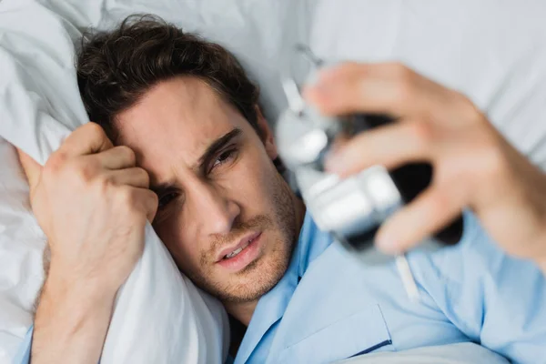 Vista de ángulo alto del hombre molesto que sostiene el reloj despertador borroso en la cama - foto de stock