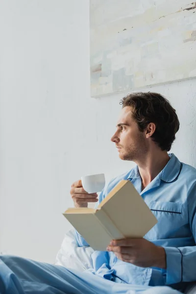 Vista laterale del giovane in pigiama con in mano una tazza di caffè e un libro sul letto — Foto stock