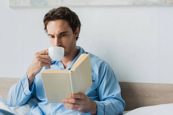Young man drinking coffee and holding blurred book on bed in morning — Stock Photo