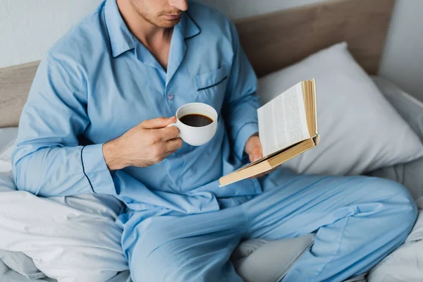 Vista recortada del hombre en pijama sosteniendo la taza de café mientras lee el libro en la cama - foto de stock