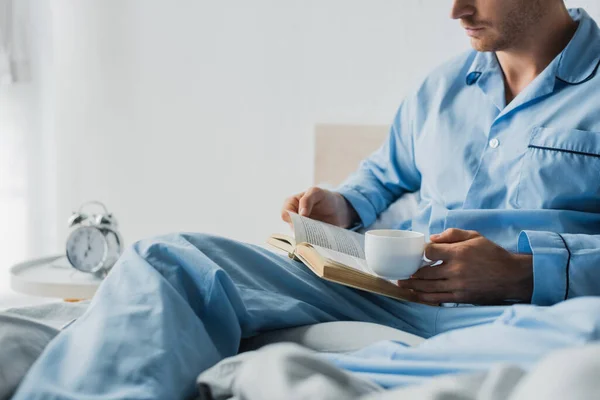 Cropped view of man in pajama holding cup and reading book near blurred alarm clock in bedroom — Stock Photo