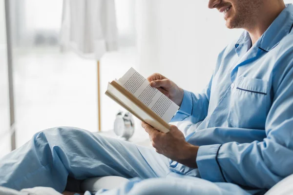 Vista recortada del hombre sonriente en pijama leyendo libro en la cama por la mañana - foto de stock