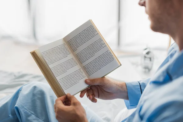 Vista ritagliata del libro nelle mani dell'uomo sfocato in pigiama sul letto — Foto stock