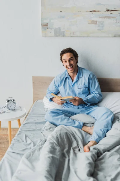 Positive man in pajama holding book and looking at camera on bed — Stock Photo
