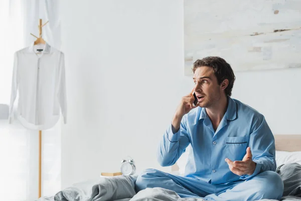 Hombre estresado en pijama hablando por teléfono celular en la cama por la mañana - foto de stock