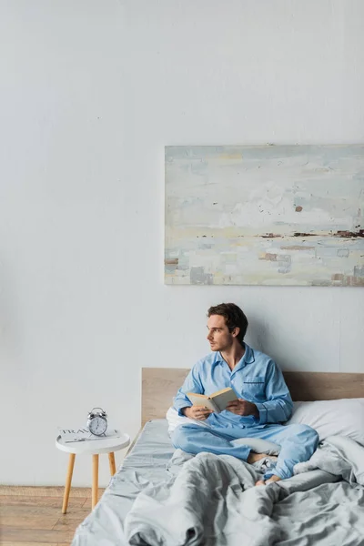 Joven en pijama sosteniendo libro cerca del despertador en la cama por la mañana - foto de stock