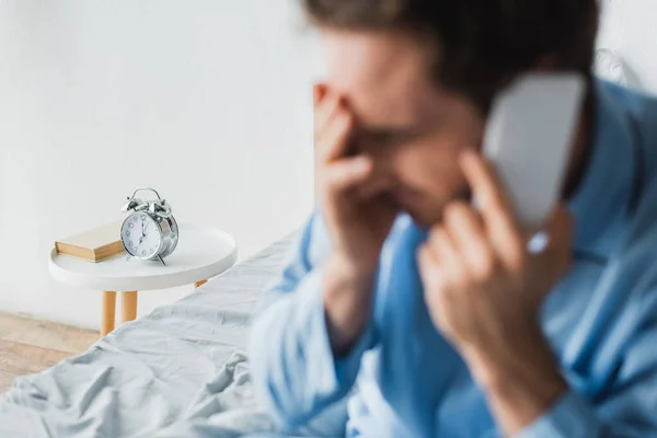 Reloj despertador en la mesita de noche cerca del hombre borroso hablando en el teléfono inteligente en el dormitorio - foto de stock