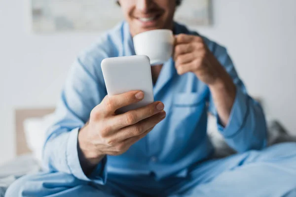Vista recortada del teléfono inteligente en la mano del hombre borroso con taza en el dormitorio - foto de stock