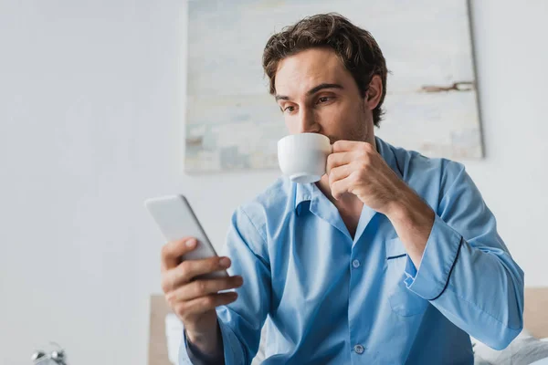 Uomo in pigiama bere caffè e utilizzando smartphone sul letto — Foto stock