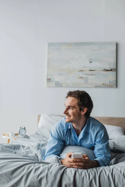 Hombre sonriente en pijama usando el teléfono celular mientras está acostado en la cama por la mañana - foto de stock