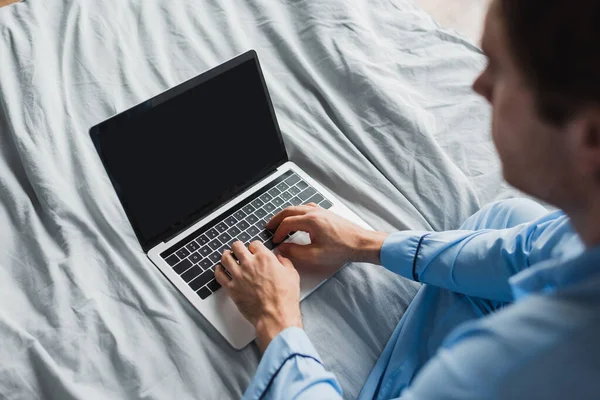 Vista de ángulo alto del hombre borroso en pijama usando el ordenador portátil con la pantalla en blanco en la cama - foto de stock