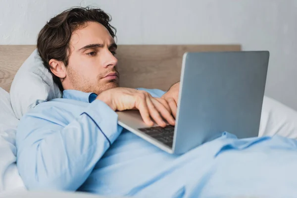 Joven freelancer en pijama usando una computadora portátil borrosa mientras está acostado en la cama por la mañana - foto de stock