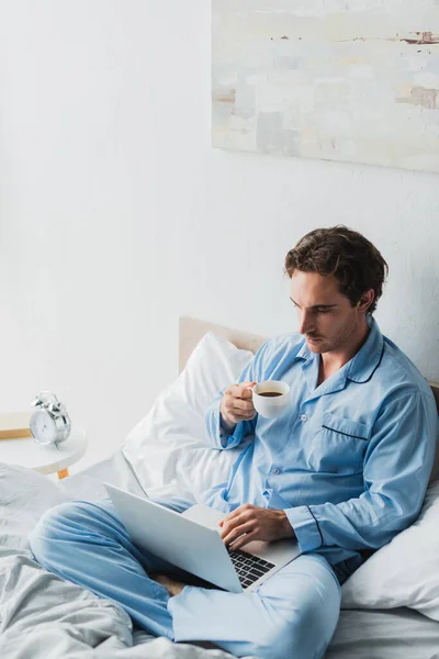 Freelancer en pijama sosteniendo una taza de café mientras usa el portátil en la cama - foto de stock