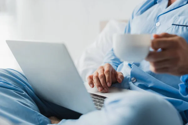 Vista recortada del freelancer en pijama sosteniendo una taza de café y usando una computadora portátil borrosa en la cama - foto de stock