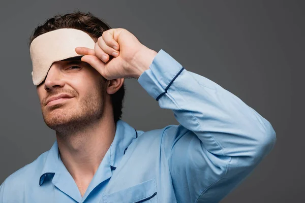 Upset man in pajama holding sleep mask and looking at camera isolated on grey — Stock Photo
