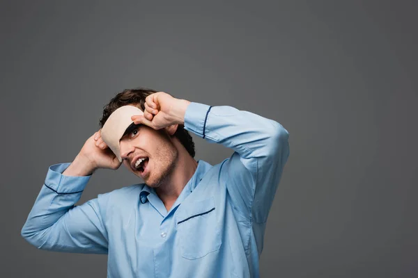 Hombre positivo en pijama sosteniendo la máscara del sueño y mirando a la cámara aislada en gris - foto de stock