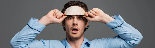 Shocked man in pajama holding sleep mask isolated on grey, banner — Stock Photo