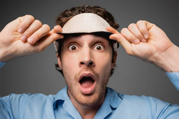 Portrait of shocked man holding sleep mask isolated on grey — Stock Photo