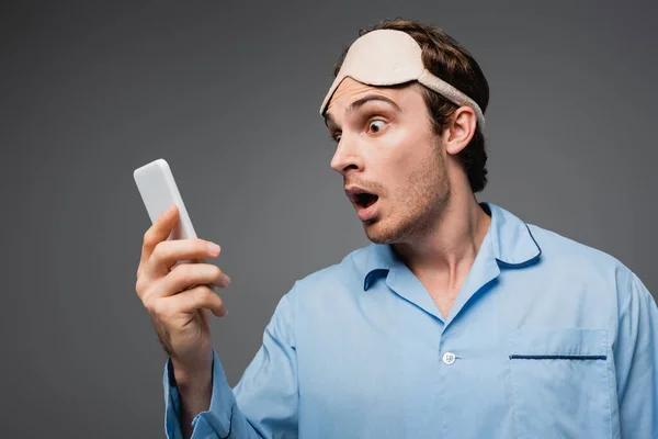 Shocked man in pajama and sleep mask looking at smartphone isolated on grey — Stock Photo