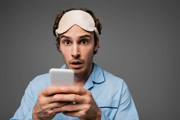 Shocked man in sleep mask and pajama holding smartphone and looking at camera isolated on grey — Stock Photo