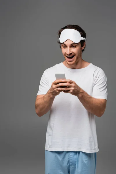 Homme positif en masque de sommeil et pyjama à l'aide d'un téléphone cellulaire isolé sur gris — Photo de stock