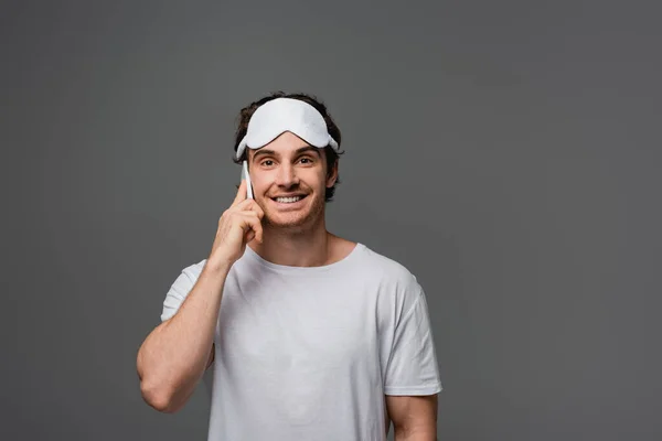 Homme souriant en masque de sommeil et t-shirt blanc parlant sur smartphone isolé sur gris — Photo de stock