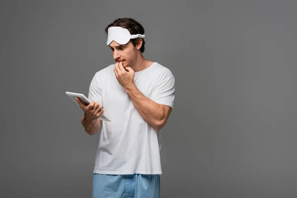 Gestresster Mann mit Schlafmaske und Pyjama blickt auf digitales Tablet isoliert auf Grau — Stockfoto