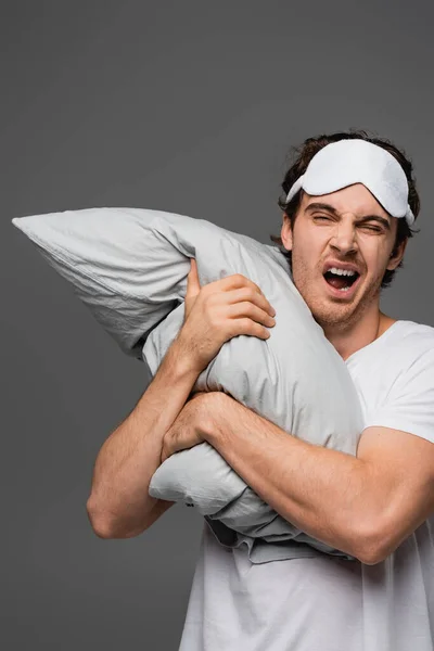 Man in sleep mask yawning while holding pillow isolated on grey — Stock Photo