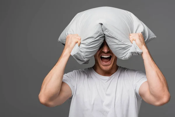 Hombre irritado en camiseta blanca gritando y sosteniendo almohada cerca de la cabeza aislada en gris - foto de stock