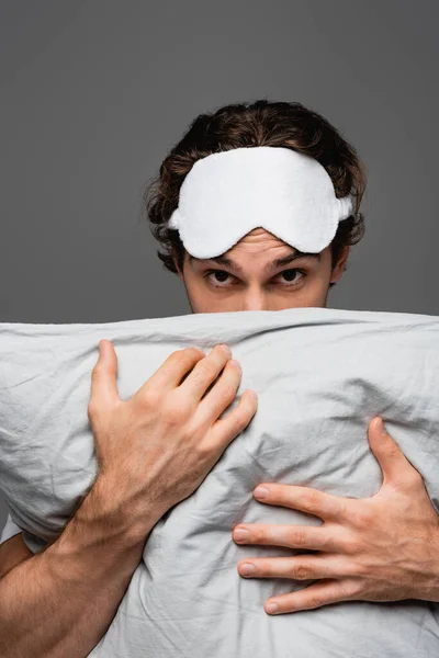 Hombre en la máscara del sueño cubriendo la cara con almohada y mirando a la cámara aislada en gris - foto de stock