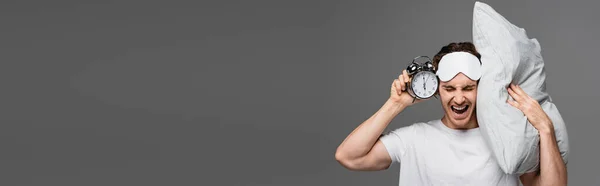 Angry man in sleep mask holding alarm clock and pillow isolated on grey, banner — Stock Photo