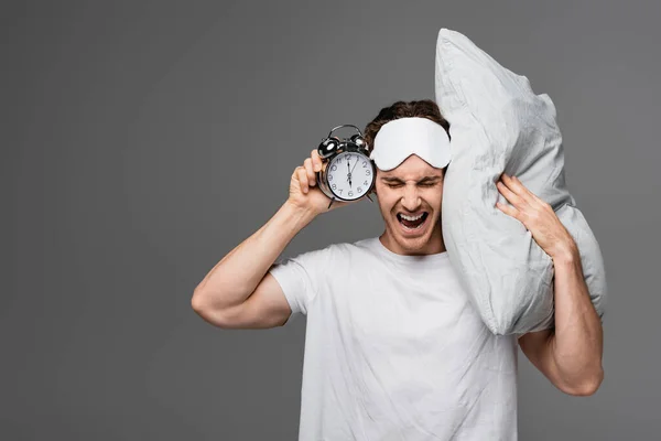 Homem irritado em máscara de sono segurando travesseiro e despertador isolado em cinza — Fotografia de Stock