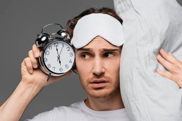 Hombre joven con máscara de sueño sosteniendo almohada y despertador aislado en gris - foto de stock