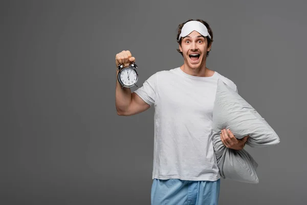 Homem feliz em pijama e máscara de sono segurando travesseiro e despertador isolado em cinza — Fotografia de Stock