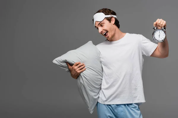 Homme positif dans le masque de sommeil tenant oreiller et réveil isolé sur gris — Photo de stock