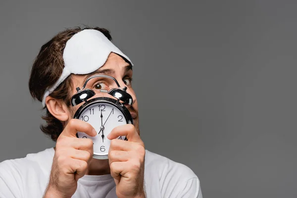 Young man in sleep mask covering face with alarm clock isolated on grey — Stock Photo